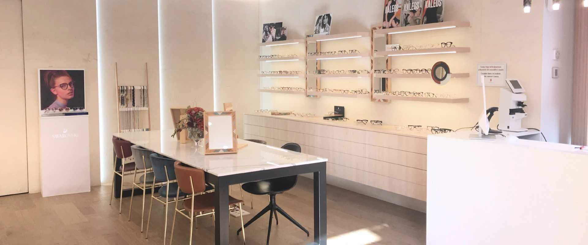 Óptica con vitrinas iluminadas de gafas y mostrador junto a sillas tapizadas de colores