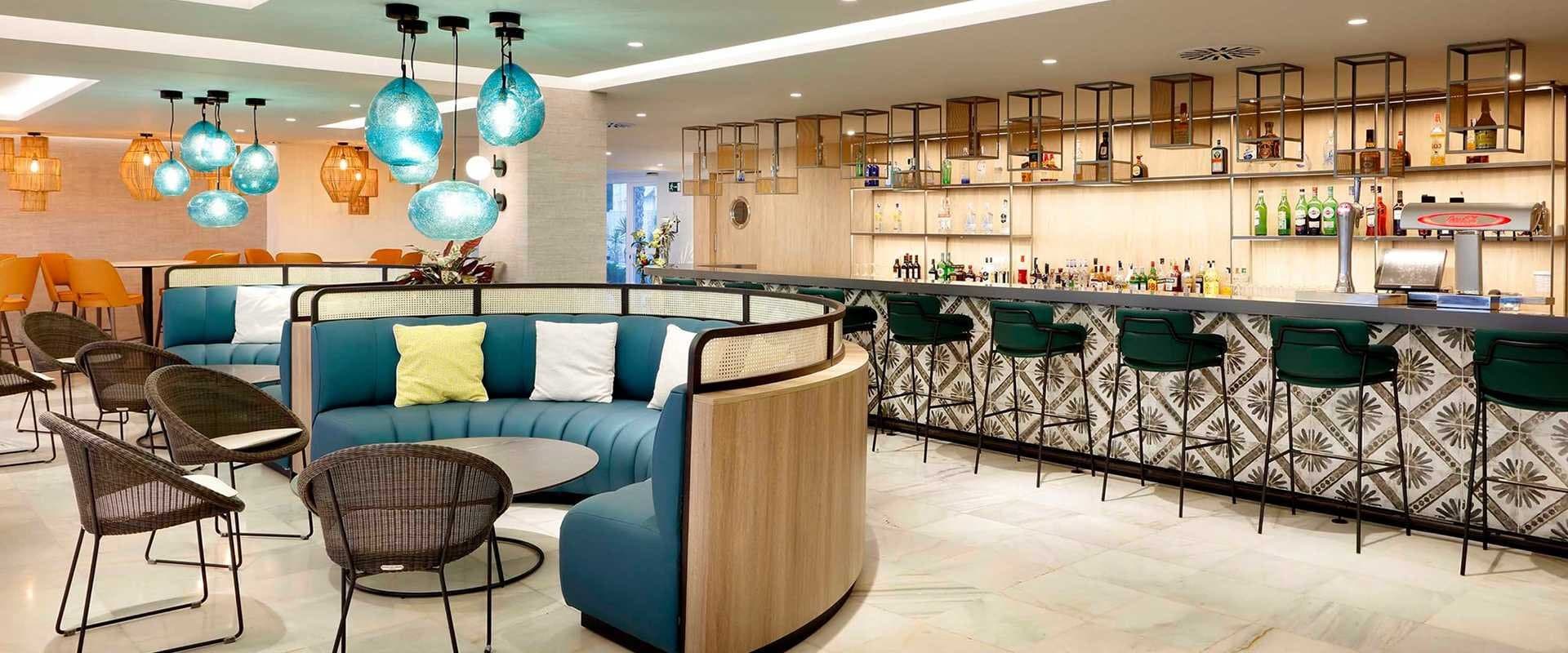 Bar de hotel con sofás azules, taburetes verdes y estanterías de botellas iluminadas