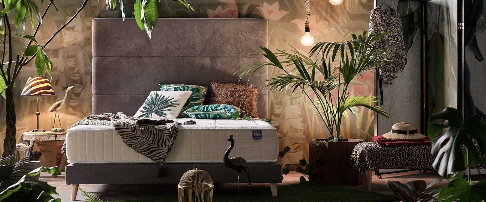 Dormitorio con cabecero tapizado gris, cojines con motivos tropicales y plantas decorativas