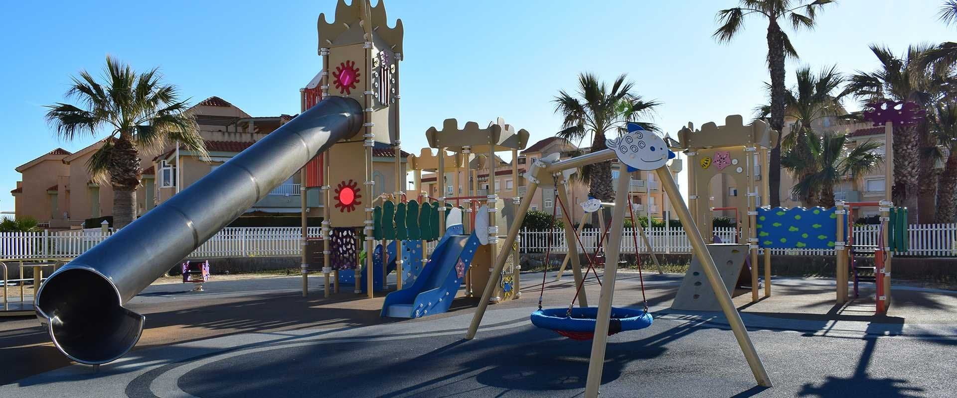 Parque infantil con tobogán metálico, columpios y torres de juego temáticas entre palmeras