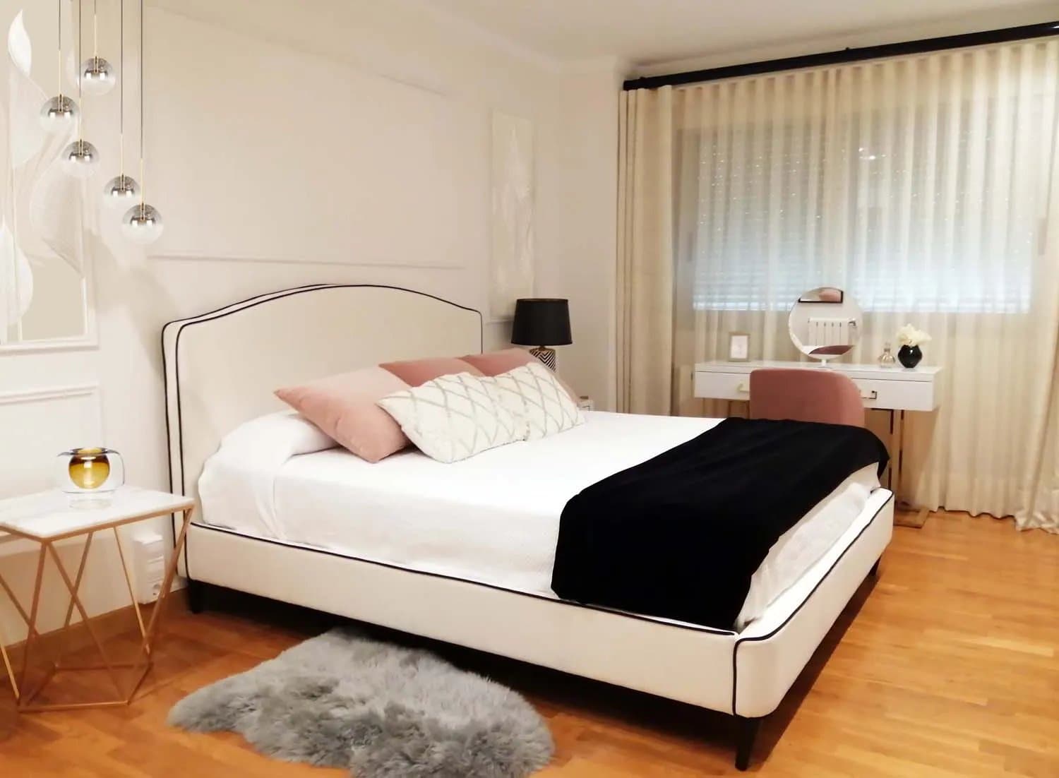 Dormitorio con cama de cabecero tapizado beige, cojines rosas y mesa auxiliar dorada