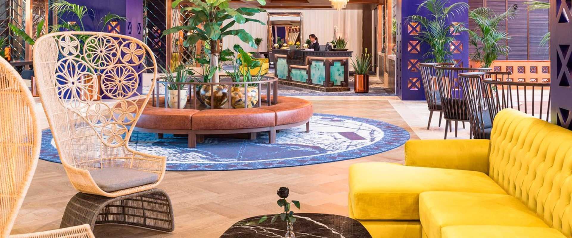Lobby de hotel con sillón de ratán, sofá amarillo y mostrador de recepción en tonos dorados