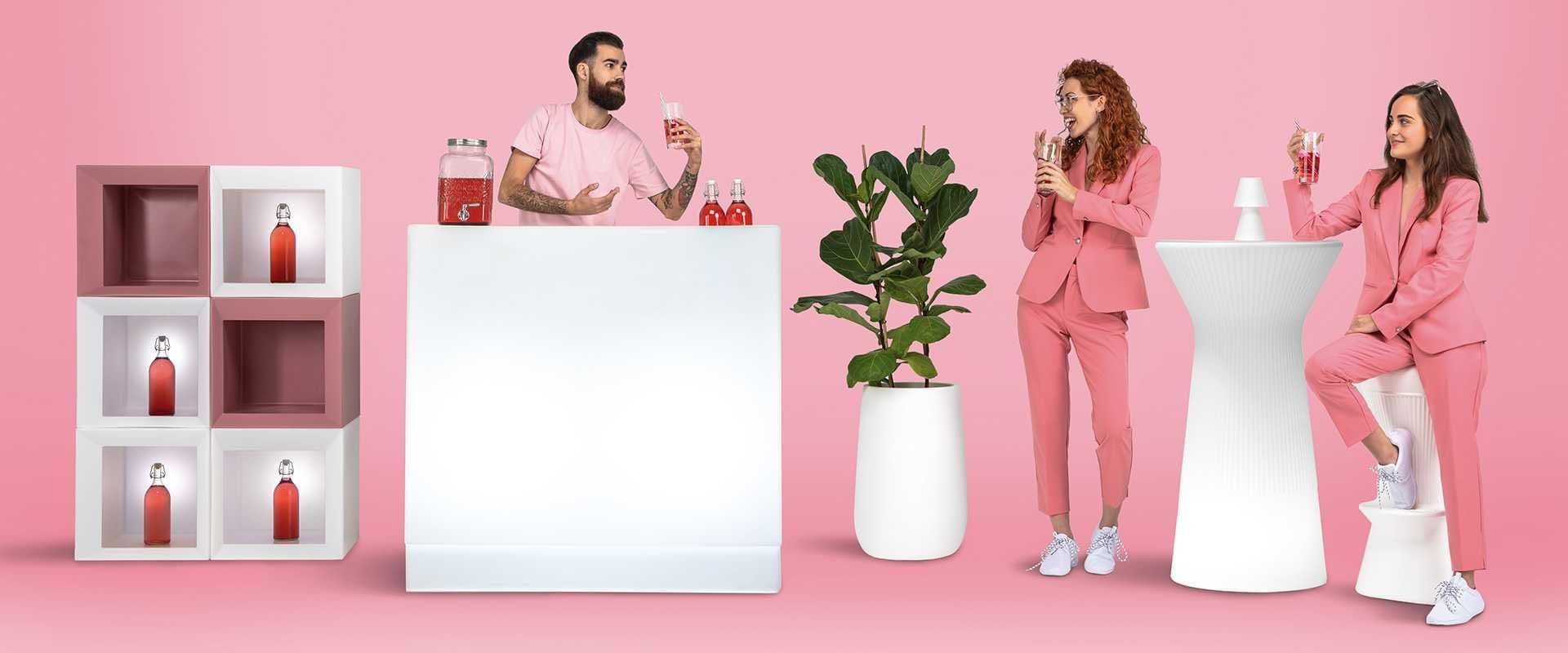 Estantería y mostrador de mimbre con botellas sobre fondo rosa, de la marca New Garden