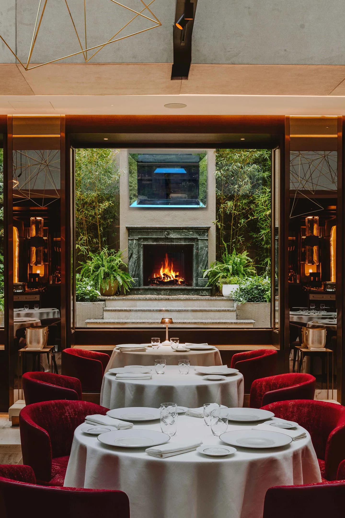 Sala principal del restaurante Saddle con mesas redondas vestidas de lino blanco, sillas de terciopelo rojo y ventanal al jardín con chimenea