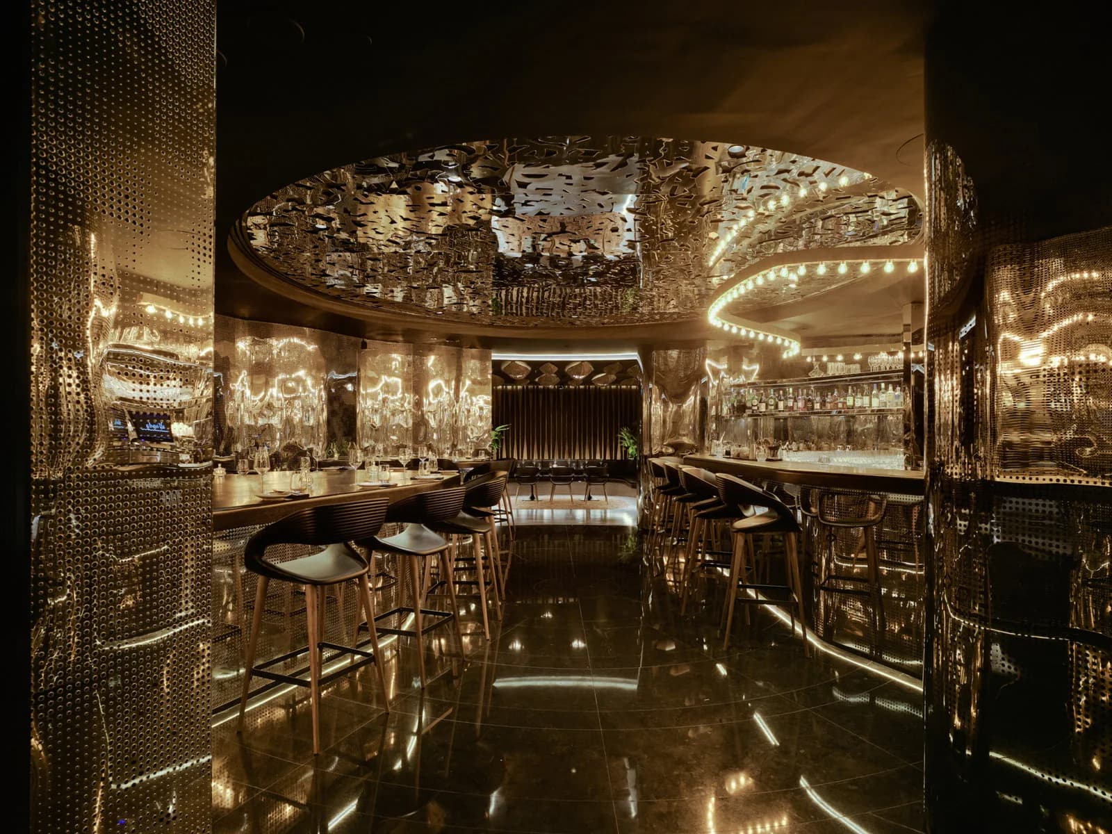 Barra de restaurante con techo de espejo dorado calado, taburetes de madera y botellas en estantería, entre paredes de espejo dorado