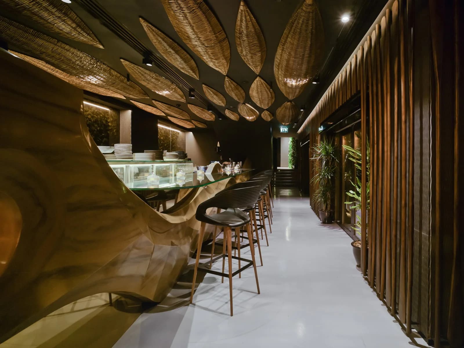 Barra de sushi dorada con taburetes negros junto a un pasillo con puertas de cristal y plantas, bajo un techo de paneles de mimbre