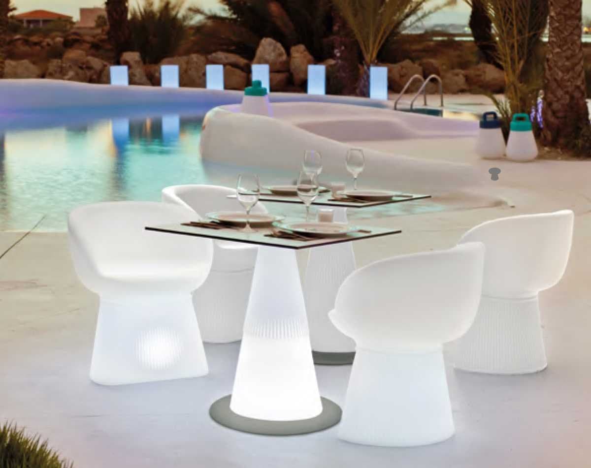 Mesa junto a una piscina iluminada al atardecer con sillas blancas luminosas y copas de cristal