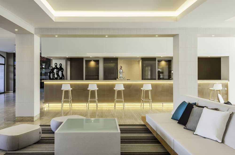 Barra de bar de hotel con taburetes blancos, estanterías iluminadas y sofá con cojines de colores