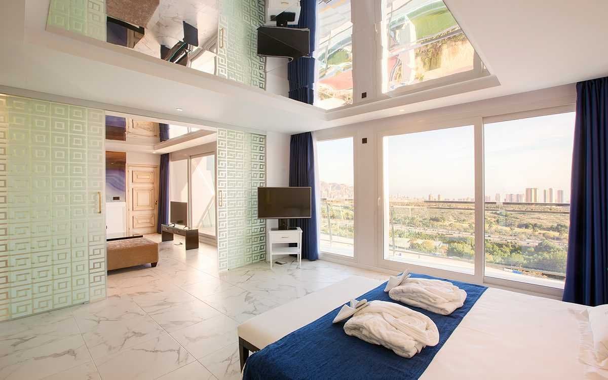 Dormitorio de hotel con cortinas azules, ventanales con vistas a la ciudad y zona de estar en altillo