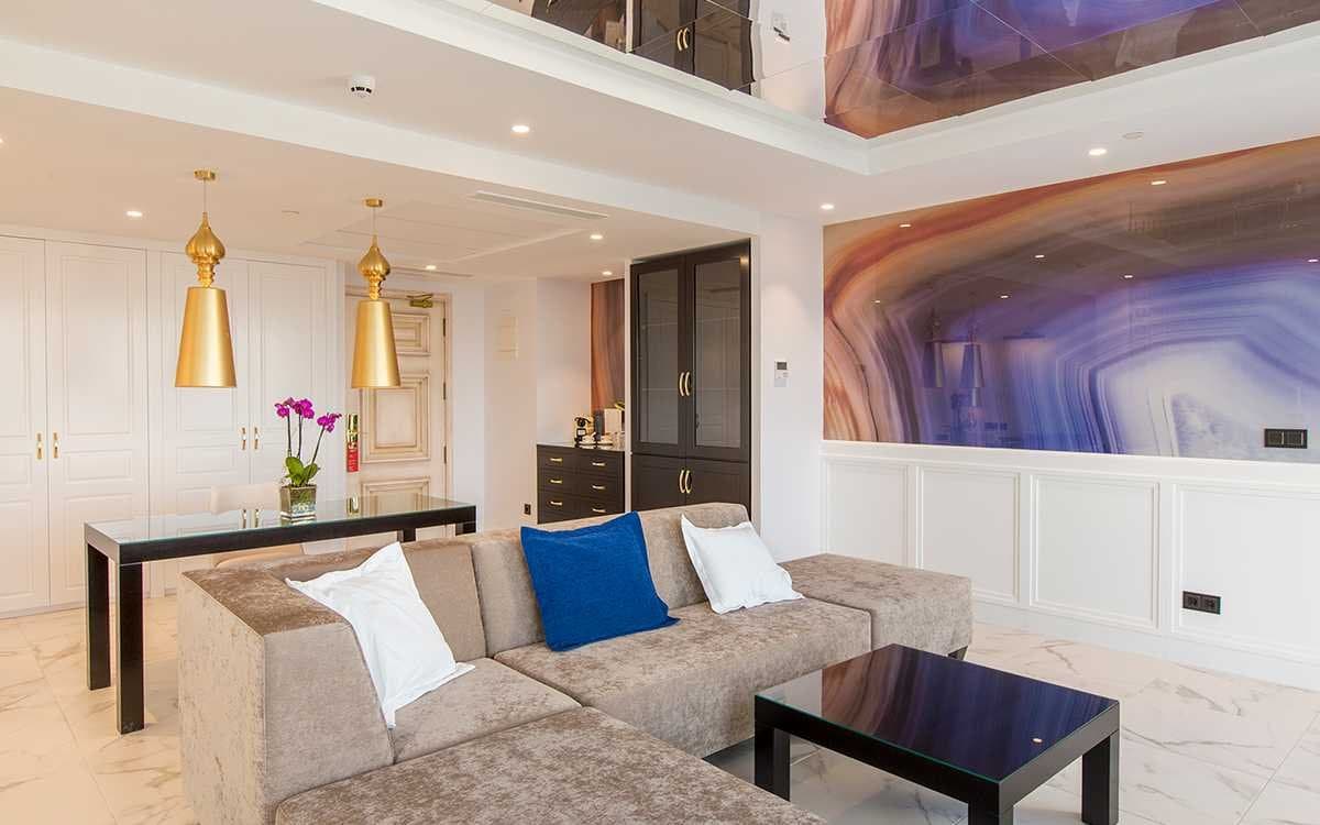 Salón de suite con sofá en tonos topo, cojín azul y pared revestida con panel de mármol veteado en tonos morados