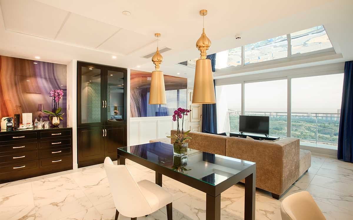 Comedor de suite con lámparas colgantes doradas, mesa de cristal y vistas panorámicas tras cortinas azules