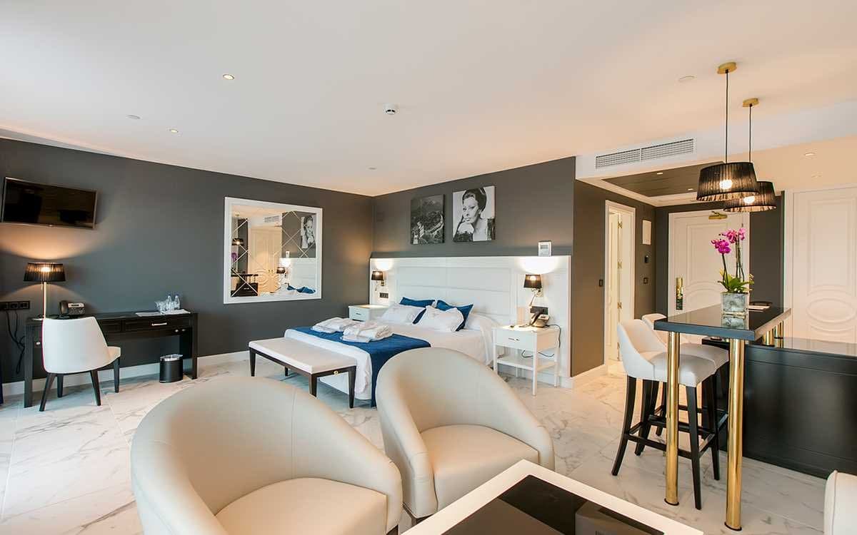 Dormitorio de hotel con cabecero tapizado blanco, sillones y zona de bar con taburetes altos