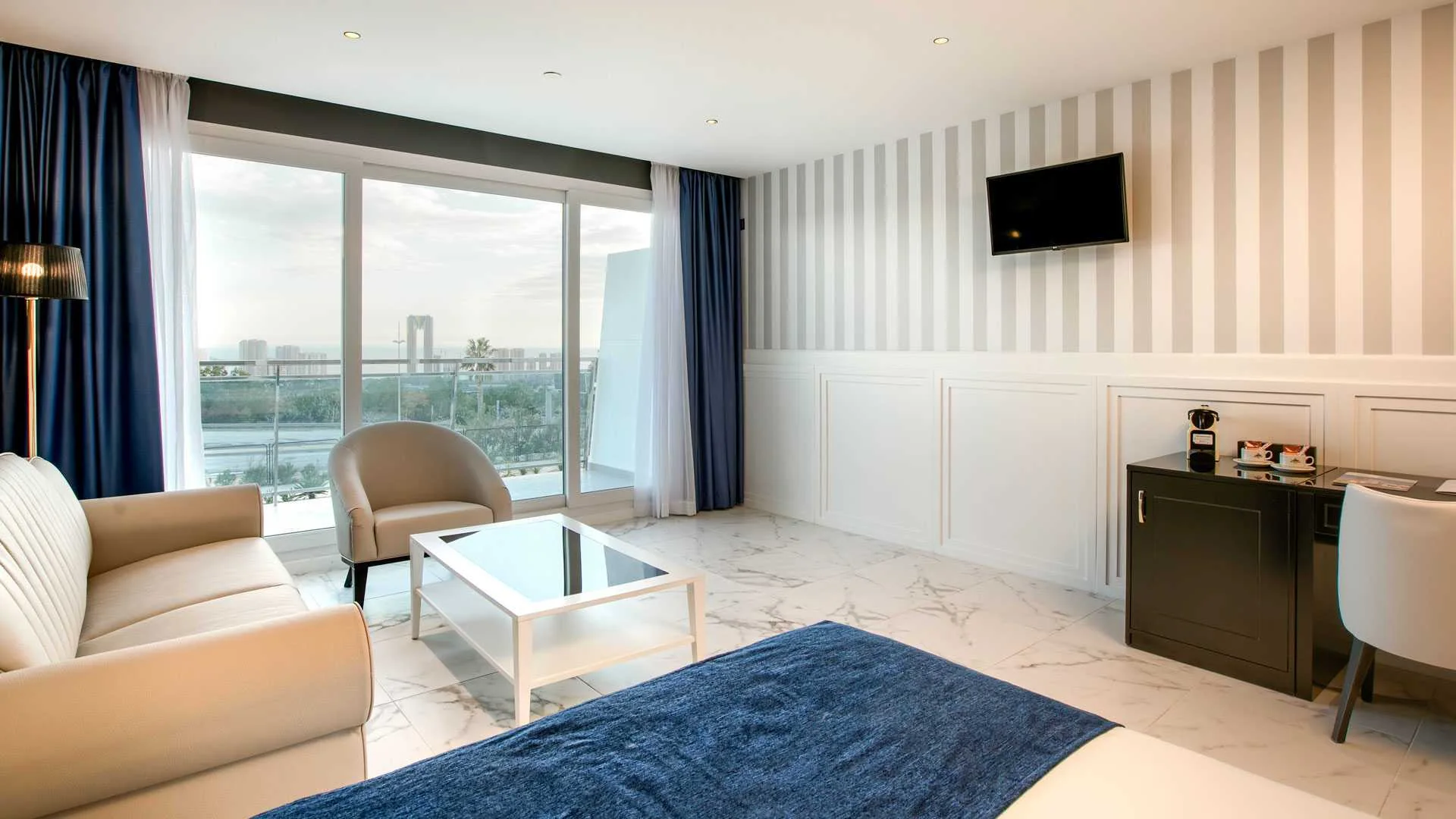 Salón de suite con sofá y sillón beige junto a un ventanal con cortinas azules, pared de rayas grises y escritorio negro