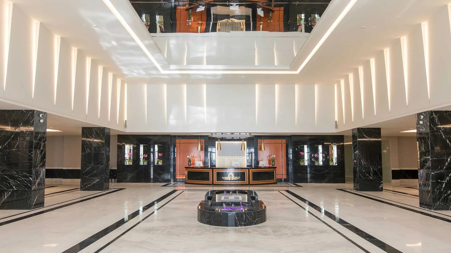 Vestíbulo amplio de hotel con columnas de mármol negro, suelo de mármol claro con filetes negros y, al fondo, la recepción con paneles naranjas