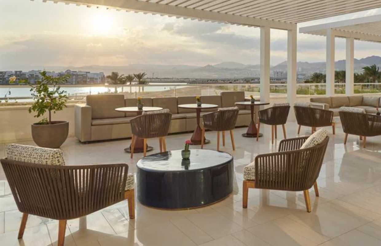 Terraza de hotel con sofás y butacas de fibra tejida junto a una mesa redonda de mármol, con vistas al mar y montañas