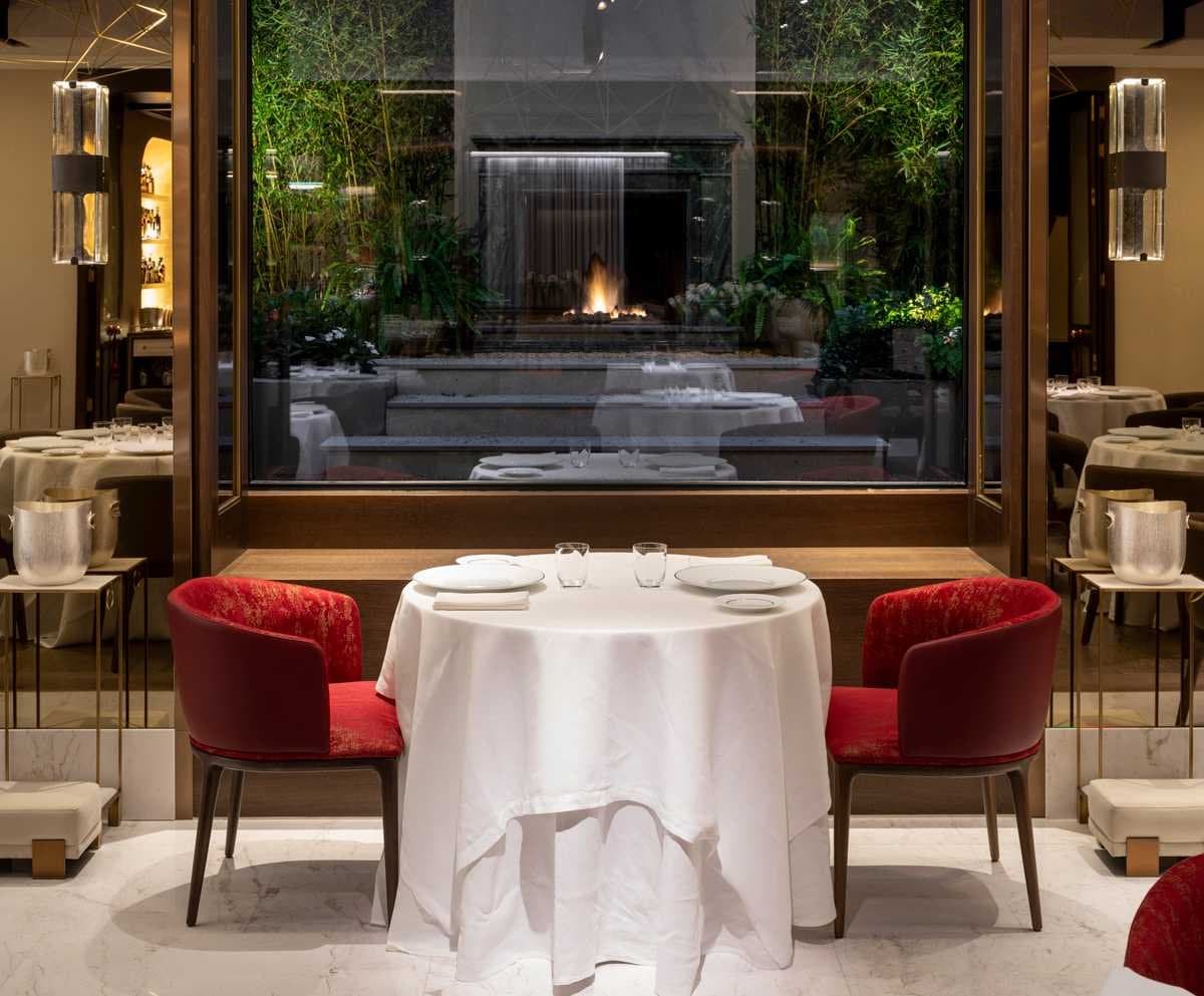 Mesa para dos junto a un ventanal con vista a un hogar de fuego y jardín de bambú, con sillas de terciopelo rojo