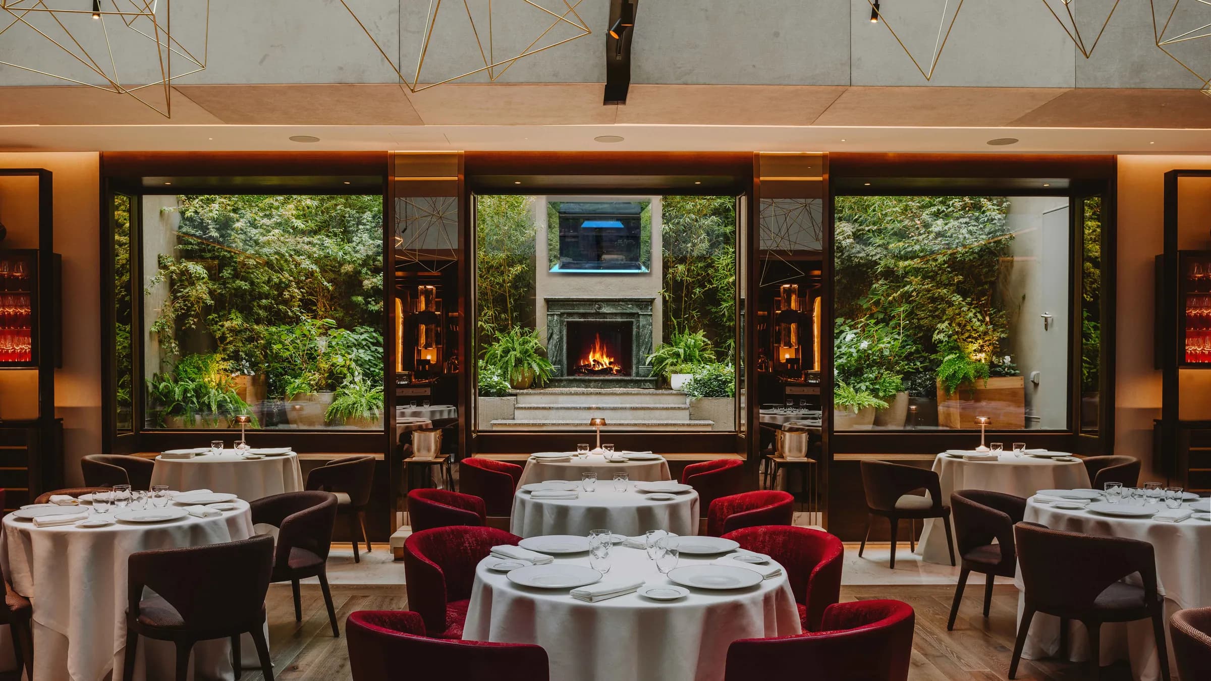 Sala principal del restaurante Saddle con mesas redondas vestidas de lino blanco, sillas de terciopelo rojo y ventanal al jardín con chimenea