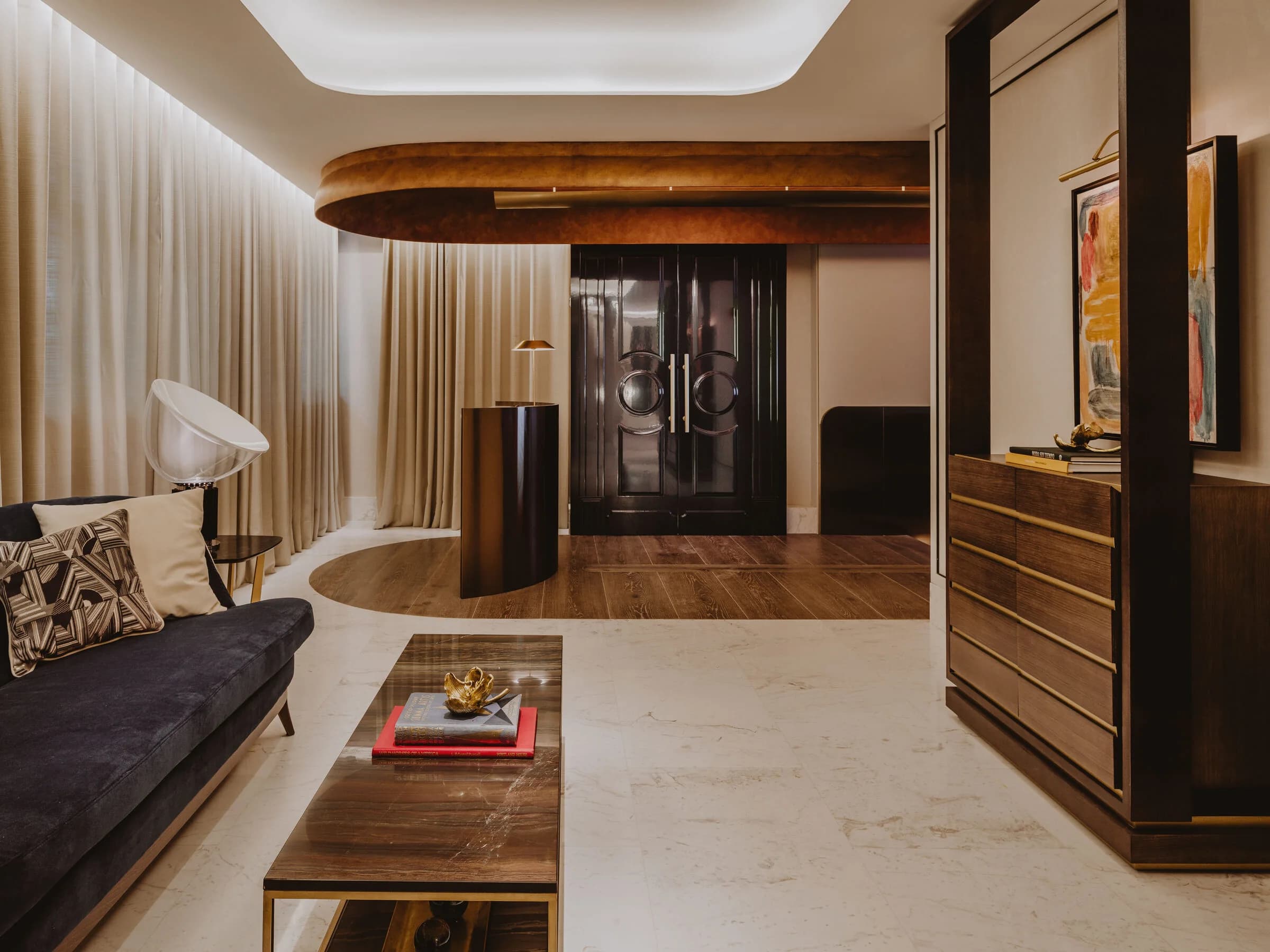 Salón de estar del restaurante con sofá oscuro, mesa baja de madera y latón, cortinas beige y una marquesina de bronce sobre una puerta lacada