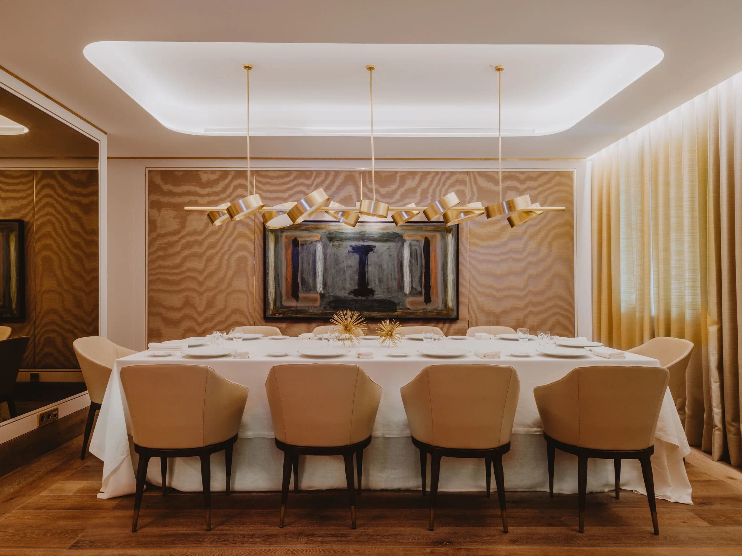 Comedor privado con pared de tejido veteado y cuadro, lámparas colgantes doradas, mesa larga con sillas tapizadas en beige y cortinas