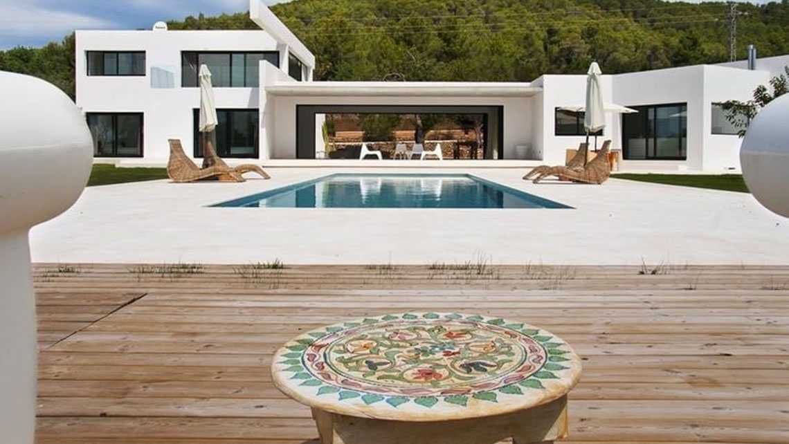 Mesa de piedra con mosaico floral en primer plano frente a una vivienda blanca con piscina y tumbonas de mimbre